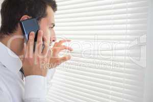 Serious businessman spying through roller blind while phoning