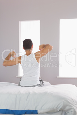 Rear view of man in pyjamas sitting on bed stretching his arms