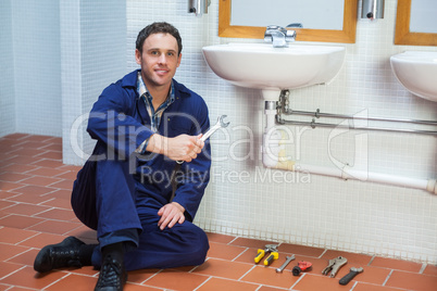 Handsome smiling plumber sitting next to sink holding wrench