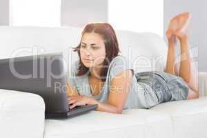 Serious young woman using her notebook while lying on her couch