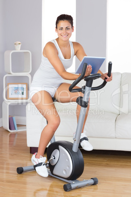 Happy training woman using an exercise bike while holding a tabl