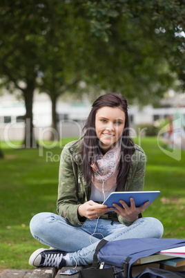 Smiling brunette student using tablet sitting on bench