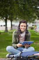 Smiling brunette student using tablet sitting on bench