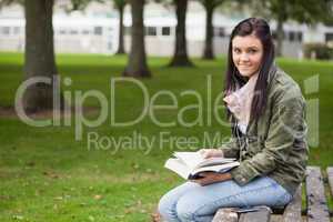 Cheerful brunette student sitting on bench reading