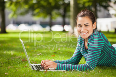 Attractive casual student lying on grass using laptop looking at