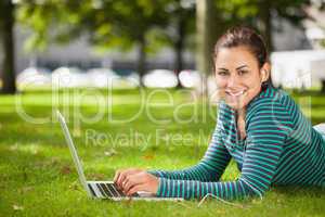 Attractive casual student lying on grass using laptop looking at