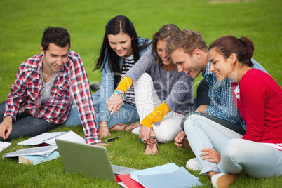 Five students sitting on the grass pointing at laptop