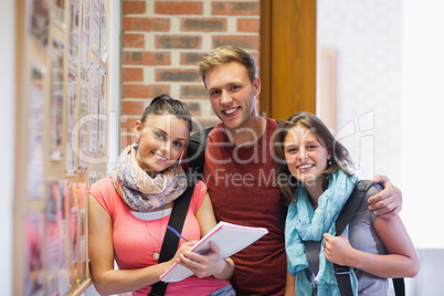 Three smiling students standing next to notice board
