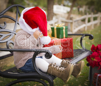 young child wearing santa hat sitting with christmas gifts outsi