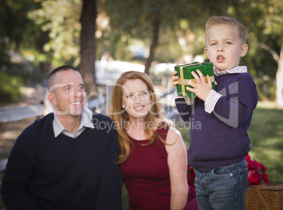 young boy holding christmas gift in park while parents look