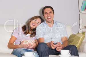 Cheerful couple watching TV with popcorn bowl on sofa at home