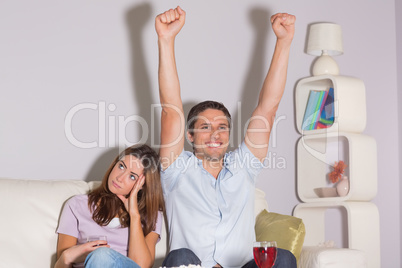 Excited man watching TV with wine by a bored woman at home
