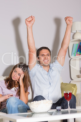 Excited man watching TV with wine and popcorn by bored woman at