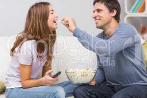Cheerful man feeding popcorn to woman on sofa