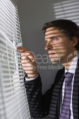 Serious young businessman peeking through blinds in office