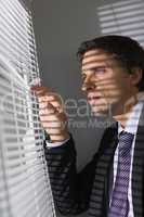 Serious young businessman peeking through blinds in office