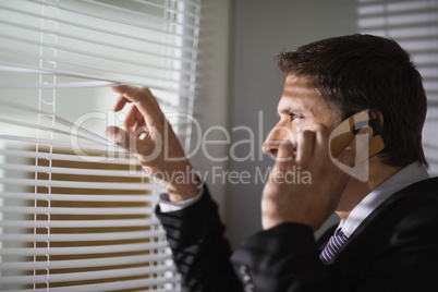 Businessman peeking through blinds while on call in office