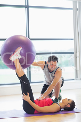 Trainer helping young woman with fitness ball at gym