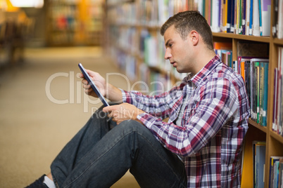 Thoughtful young student sitting on library floor using tablet