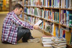 Handsome young student sitting on library floor reading book