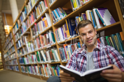 Smiling young student sitting on library floor reading book