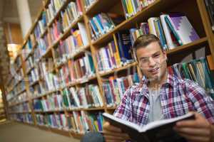 Smiling young student sitting on library floor reading book