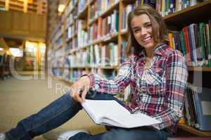 Smiling pretty student sitting on library floor reading book