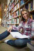 Smiling pretty student sitting on library floor holding book