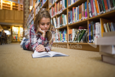 Pretty focused student lying on library floor reading book