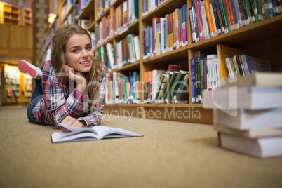 Pretty cheerful student lying on library floor reading book