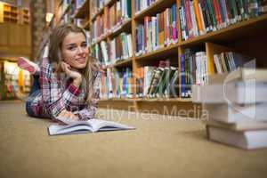 Pretty cheerful student lying on library floor reading book
