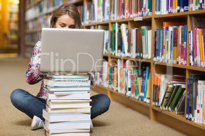 Young student sitting on library floor using laptop on pile of b