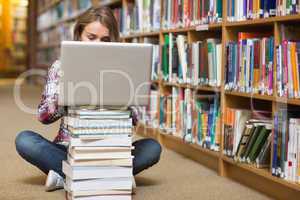 Young student sitting on library floor using laptop on pile of b