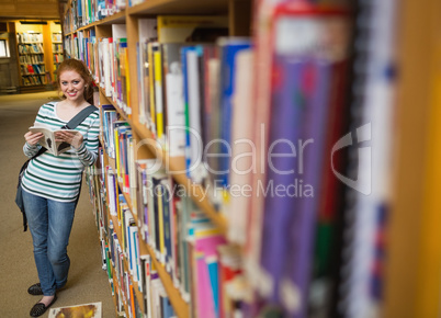 Cheerful student reading book leaning on shelf in library