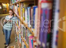 Cheerful student reading book leaning on shelf in library