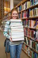 Cheerful student holding huge pile of books standing in library