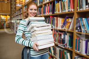 Smiling student holding heavy pile of books standing in library