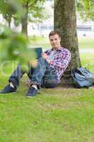 Smiling student using his tablet pc outside leaning on tree