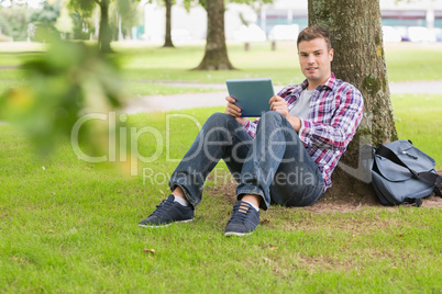 Happy student using his tablet pc outside leaning on tree