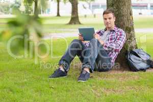Happy student using his tablet pc outside leaning on tree
