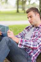 Serious student using his tablet pc outside leaning on tree