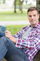 Cheerful student using his tablet pc outside leaning on tree