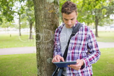 Handsome student leaning on tree using his tablet pc