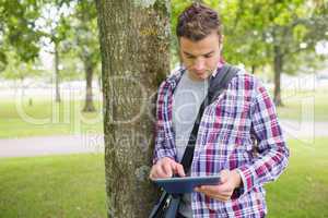 Handsome student leaning on tree using his tablet pc