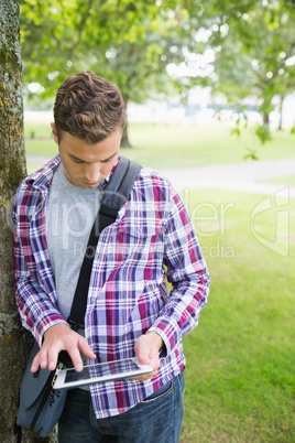 Handsome student leaning on tree using his tablet