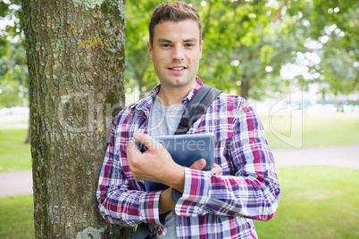 Smiling student leaning on tree holding his digital tablet