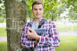 Smiling student leaning on tree holding his digital tablet