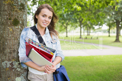 Smiling student leaning on tree holding her books