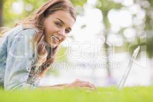 Happy student lying on the grass using her laptop looking at cam