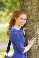 Smiling redhead student leaning on tree looking at camera